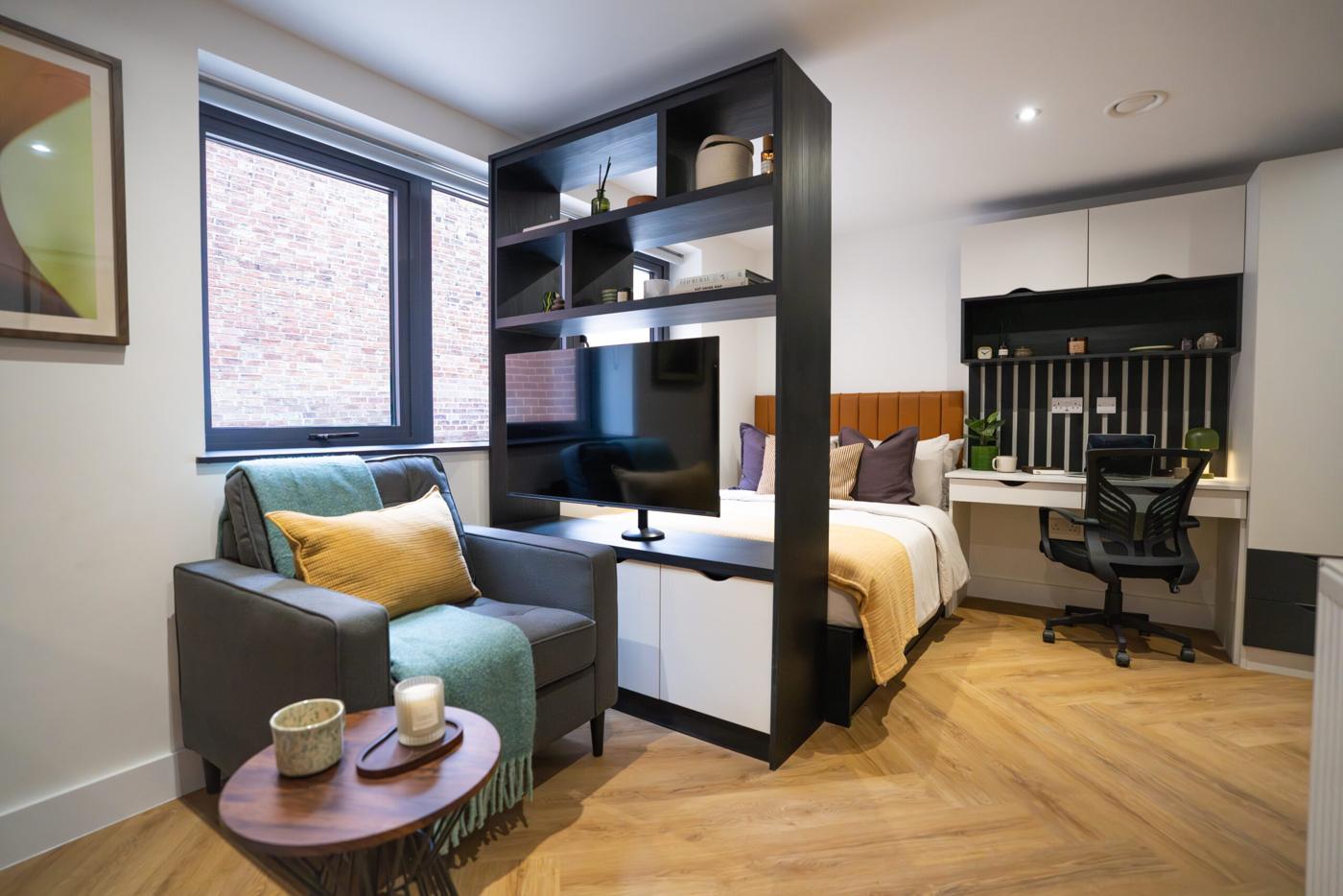 New Walk studio with a black room-divider shelving unit holding a TV, bed and desk beyond